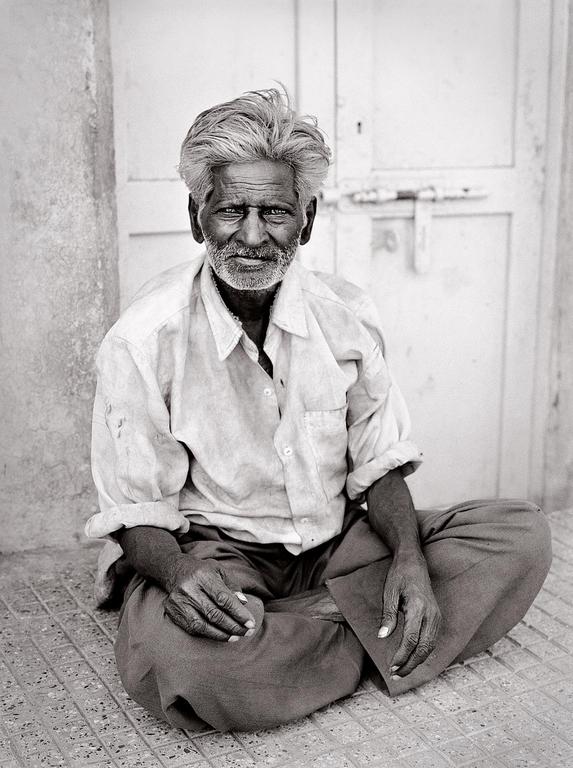 A man with gray hair and a beard sits cross-legged on a tiled surface in front of a door.