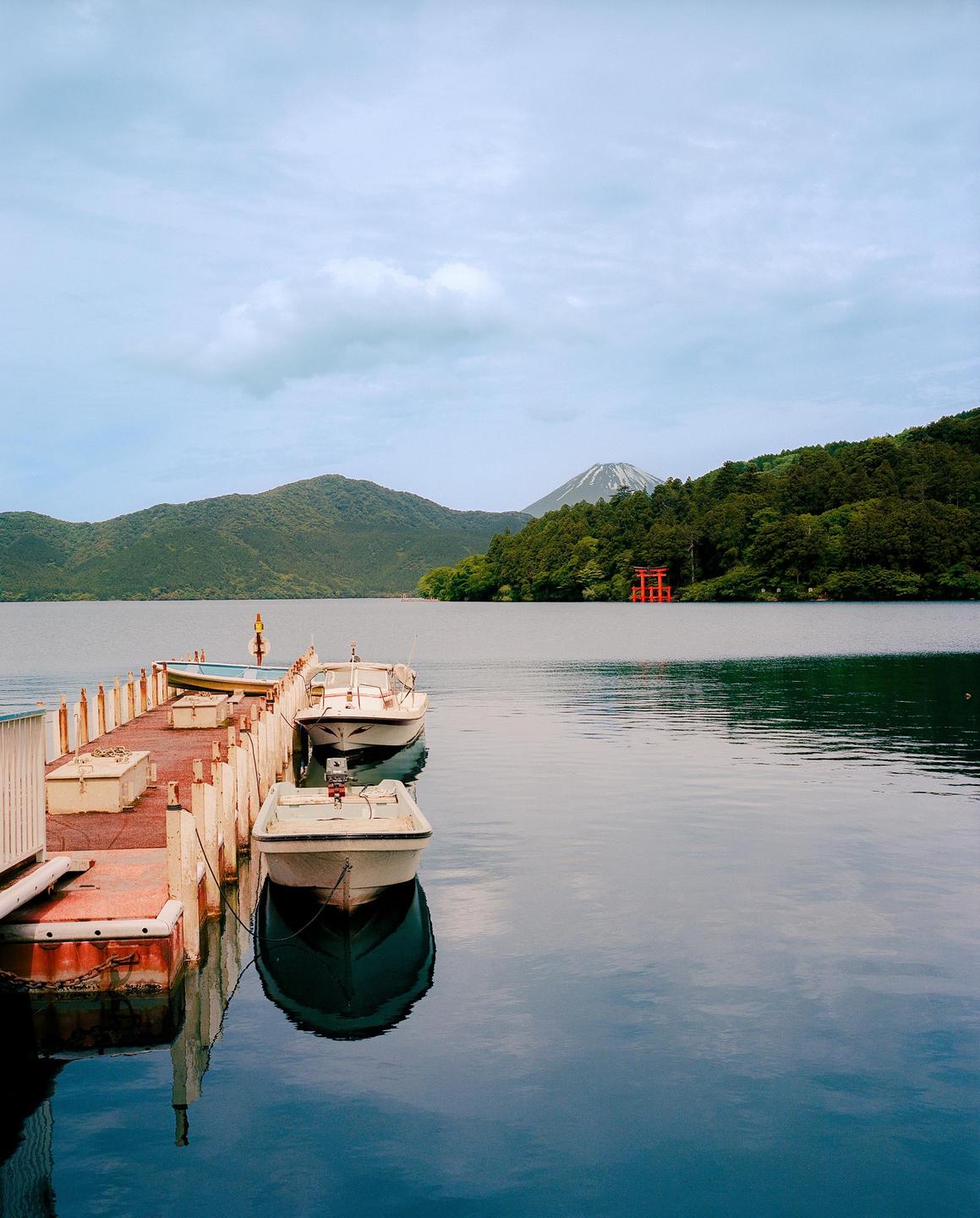 Boats are docked at a pier on a lake with a red torii gate and a snow-capped mountain in the distance.