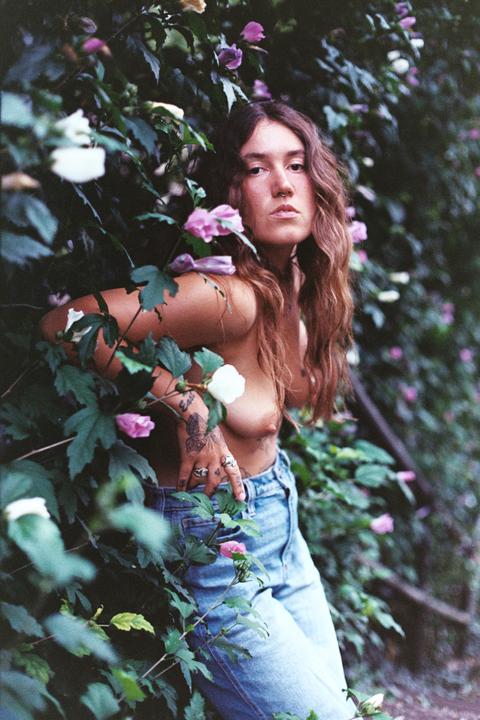 A topless woman with long hair and jeans stands among green foliage and pink and white flowers.