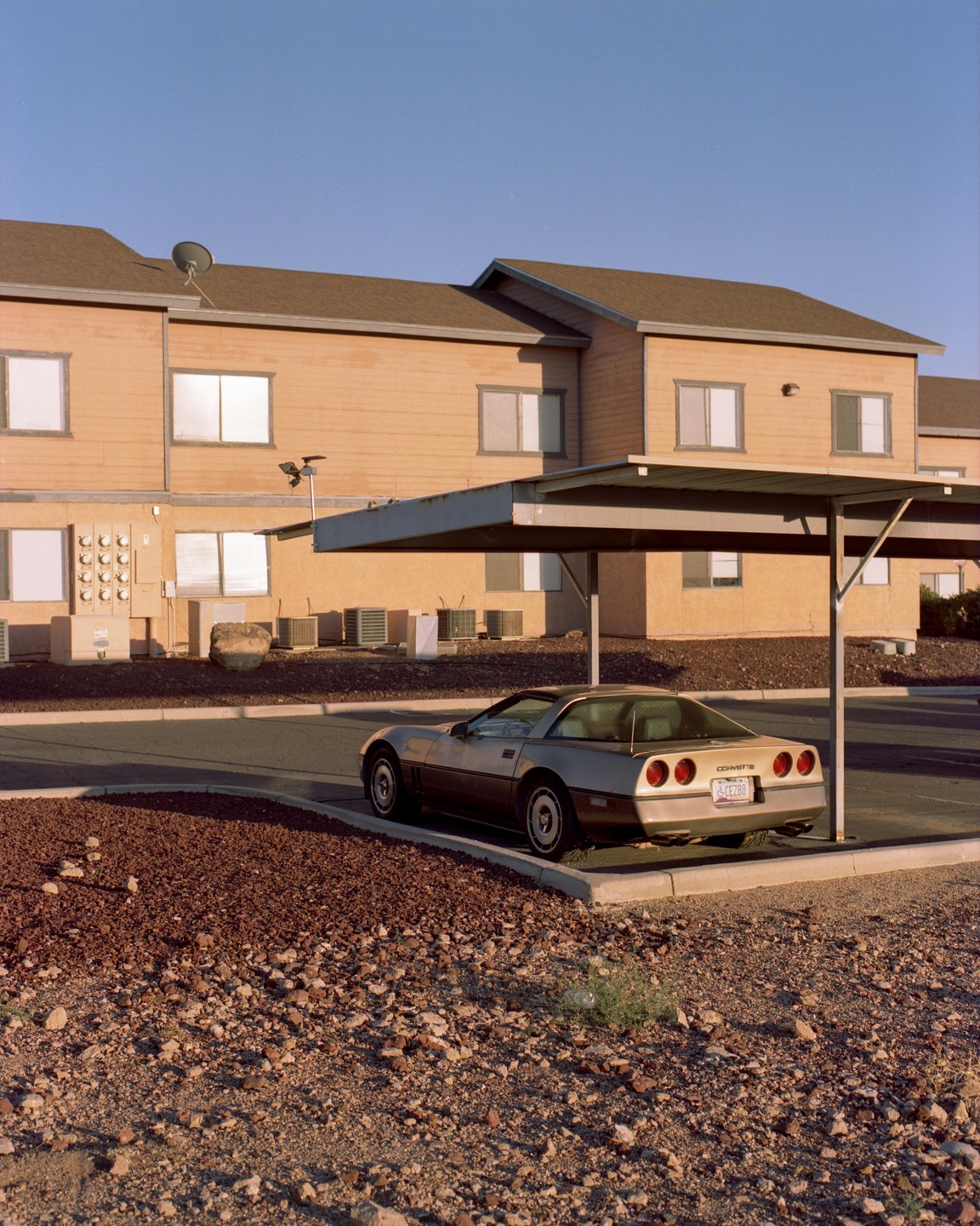 A gold corvette is parked under a carport next to a building with a gravel foreground.