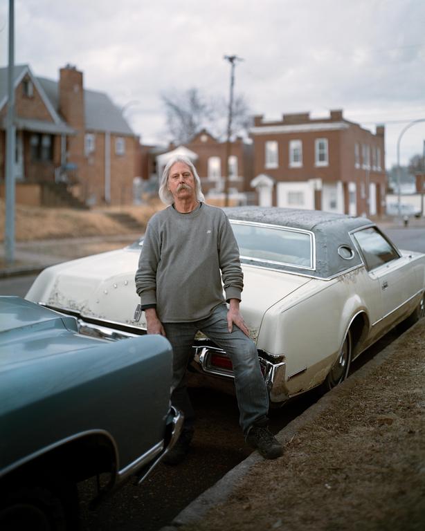 A man with long hair and a mustache stands next to a vintage car on a street with buildings in the background.