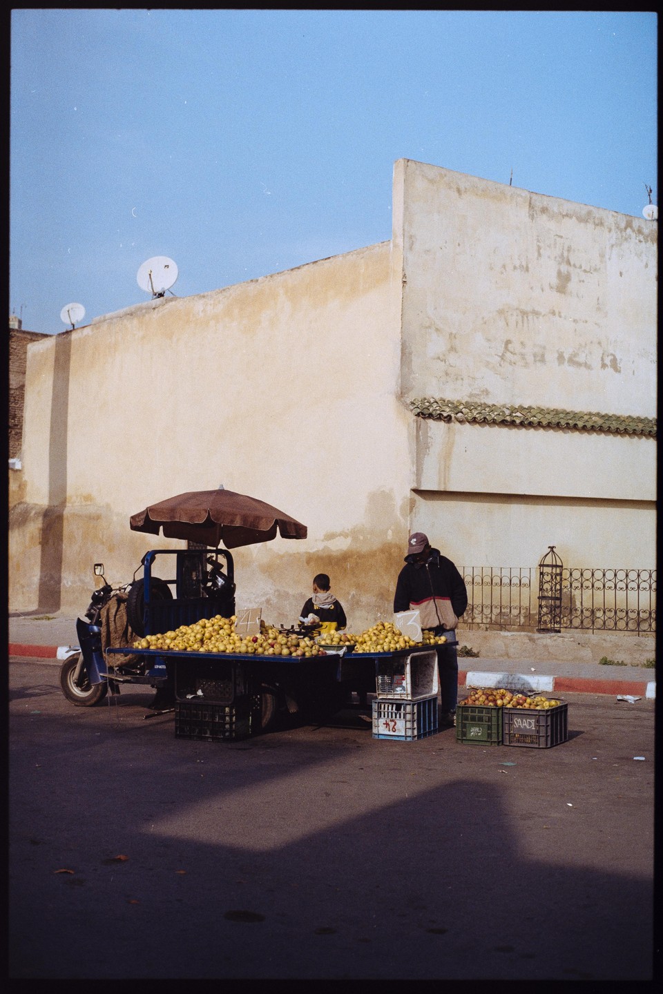 A man and a boy stand next to a fruit stand on a street with a large building in the background.