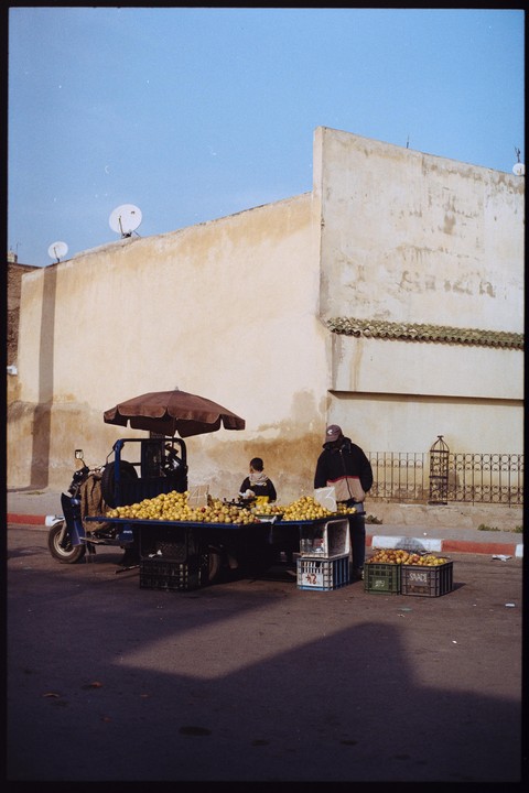 A man and a boy stand next to a fruit stand on a street with a large building in the background.
