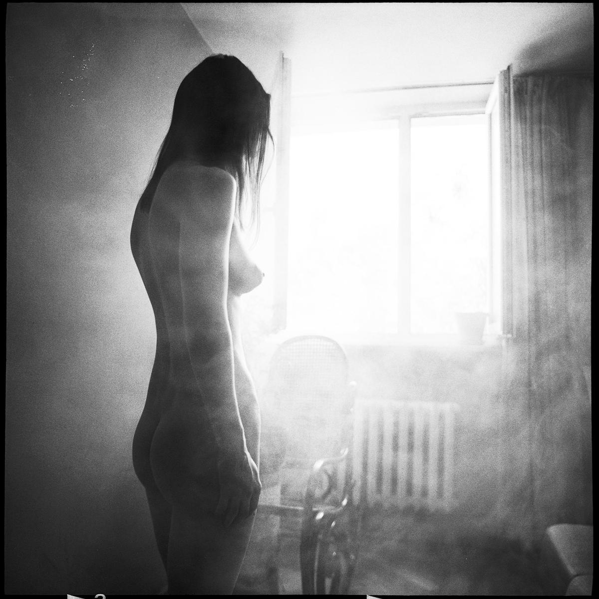 A nude woman stands in a hazy room with a bright window in the background.