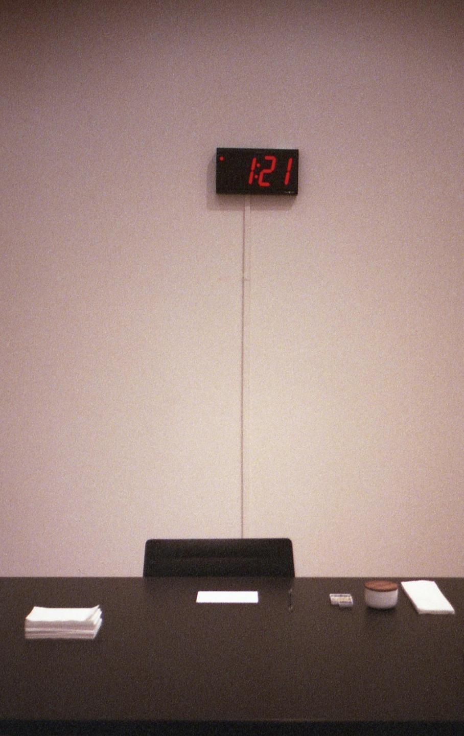 A digital clock displaying "1:21" hangs above a dark table with a chair and some items.
