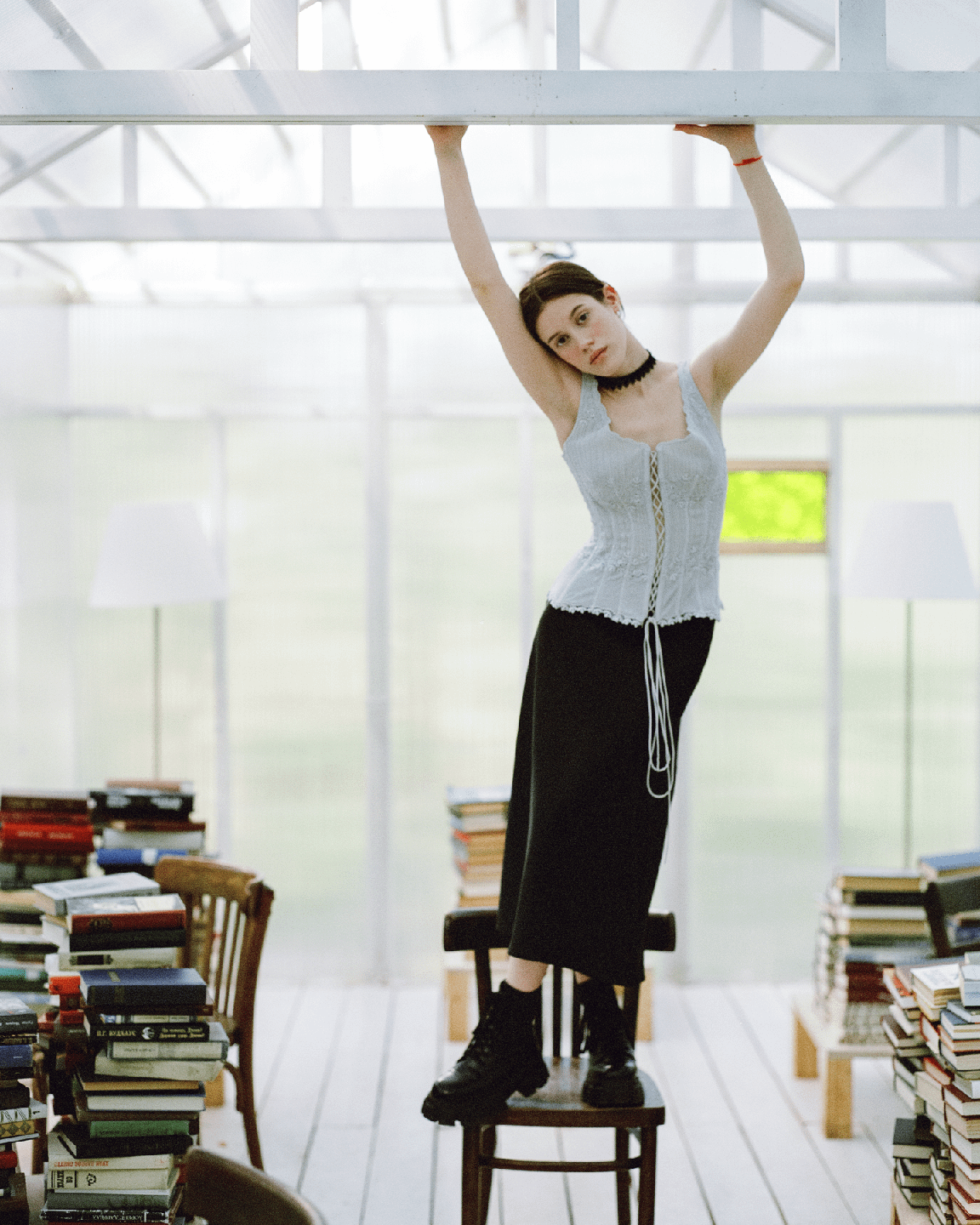 A woman stands on a chair, holding onto a white beam, surrounded by stacks of books in a bright room.