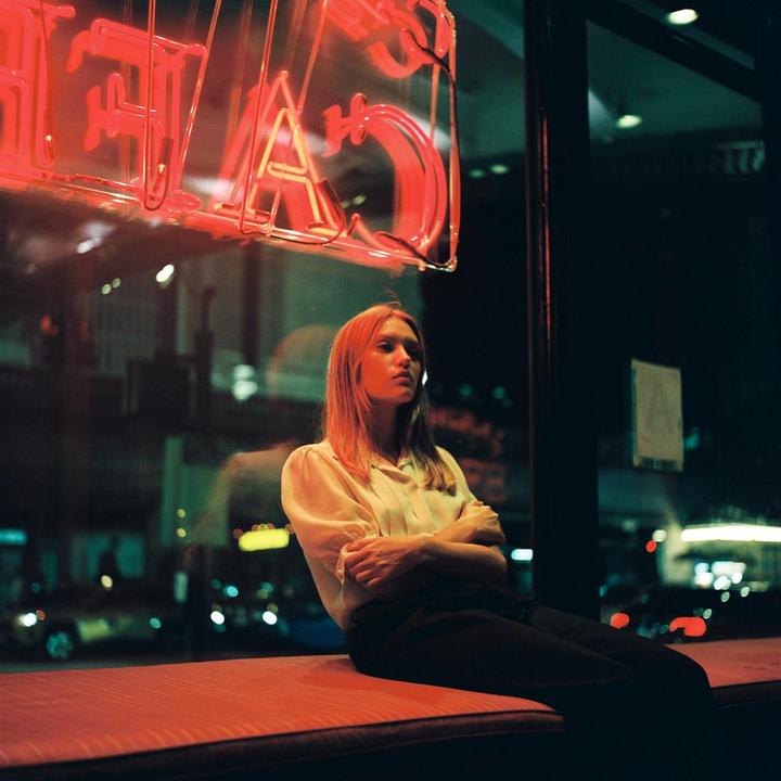 A woman sits with her arms crossed on a red bench next to a window with neon lights reflecting on the glass.
