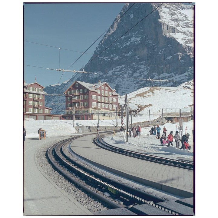 A train track curves past a hotel and people in the snow with a mountain in the background.