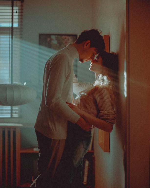 A couple stands close together, foreheads touching, with light streaming through blinds onto them.