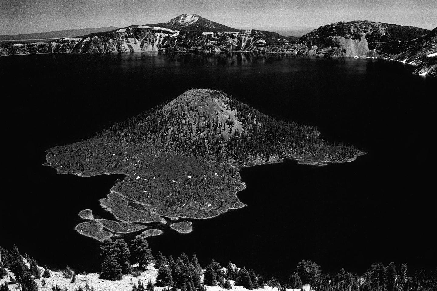 A black and white landscape shows a large lake with a tree-covered island in the middle, surrounded by mountains.