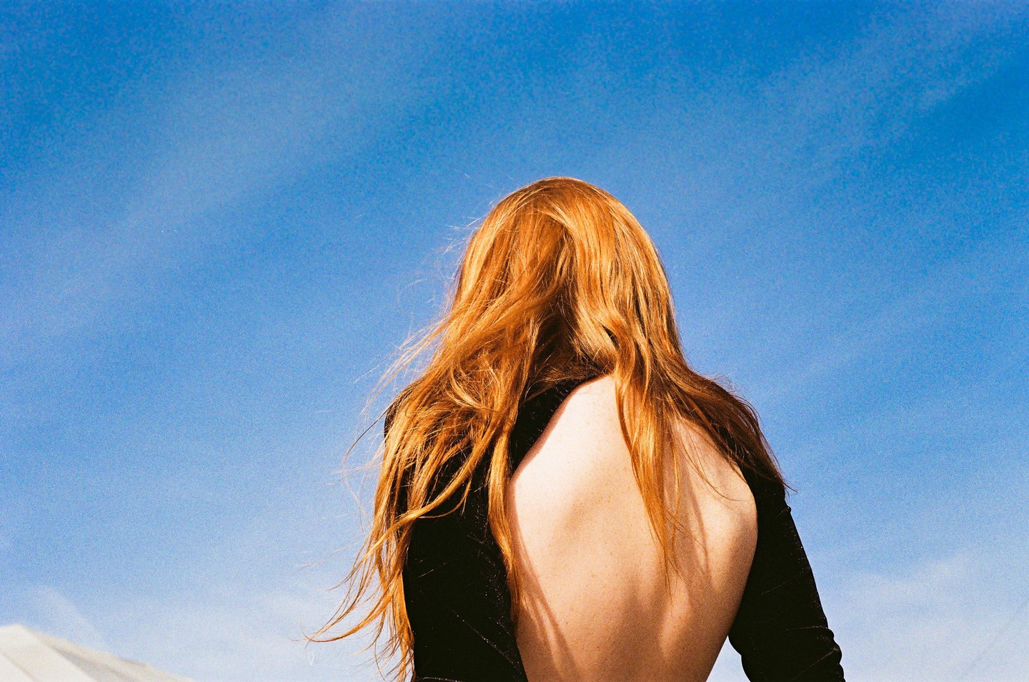 A woman with long red hair and an open-back top stands with her back to the camera against a clear blue sky.
