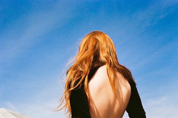 A woman with long red hair and an open-back top stands with her back to the camera against a clear blue sky.