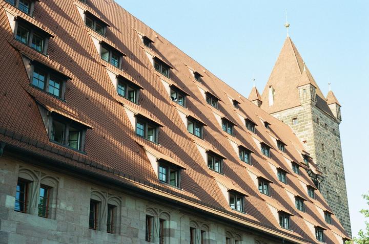 A large building with a red tile roof and many dormer windows stands next to a stone tower with a pointed roof.