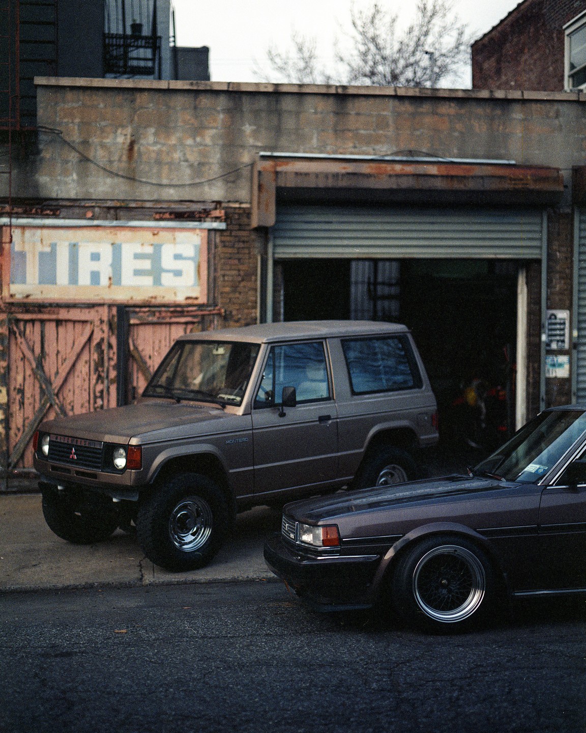 Two brown cars are parked on a street in front of a building with a "TIRES" sign.