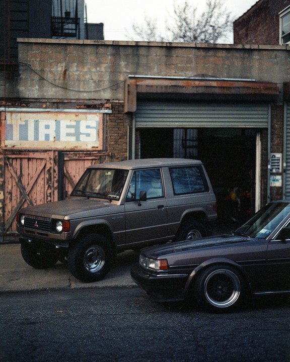 Two brown cars are parked on a street in front of a building with a "TIRES" sign.