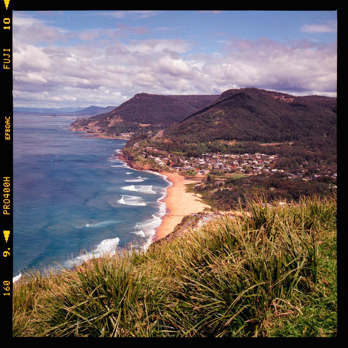 A coastal town with a beach and mountains is seen from a high vantage point with tall grass in the foreground.