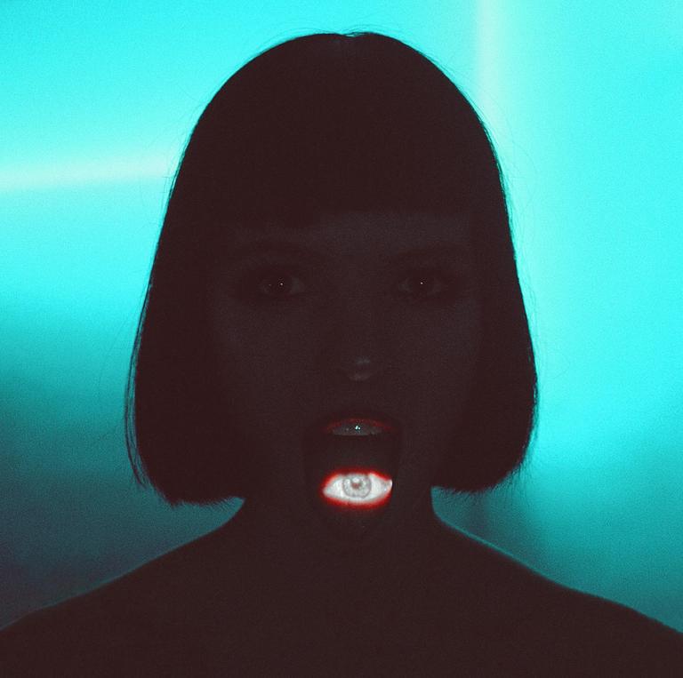 A woman with dark hair and a bob haircut has an eye projected onto her open mouth, with a bright blue background.
