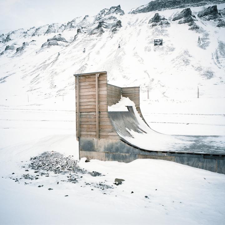 A wooden half-pipe covered in snow sits in a snowy landscape with mountains in the background.