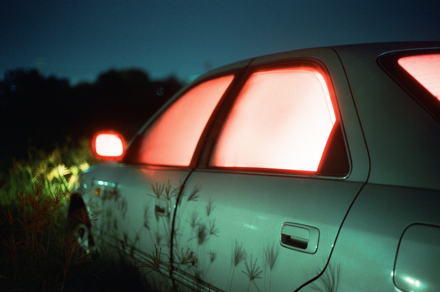 A car with glowing red windows is parked in tall grass at night.