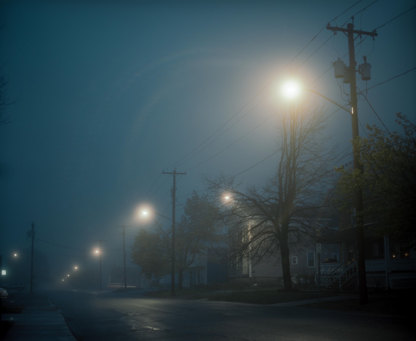 A foggy street at night is illuminated by several streetlights and the lights of a house.