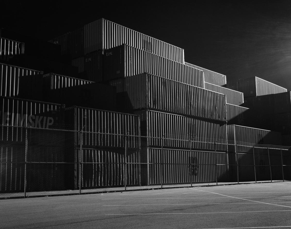 Shipping containers are stacked high behind a chain link fence at night.