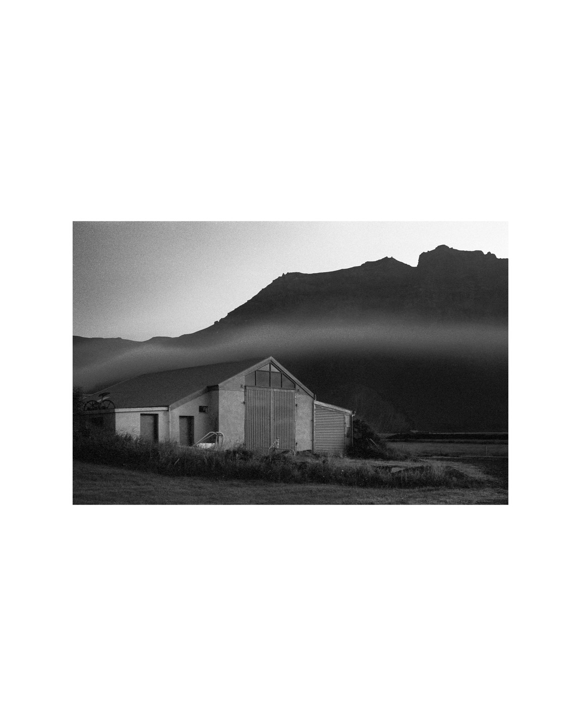A barn sits in a field with mountains in the background, partially obscured by fog.