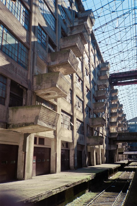 A long concrete building with many windows and small balconies stands next to train tracks under a glass roof.