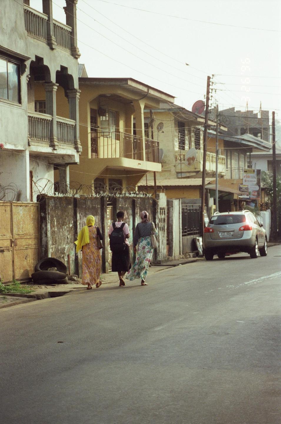Three women walk along a street past buildings and a parked car.