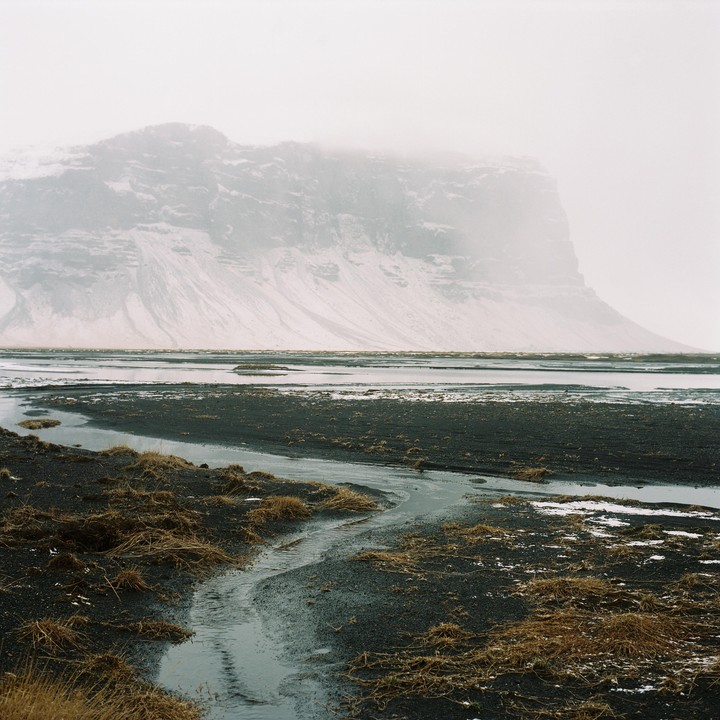 A stream winds through dark, rocky terrain with patches of dry grass, leading towards a body of water and a snow-covered mountain shrouded in mist.