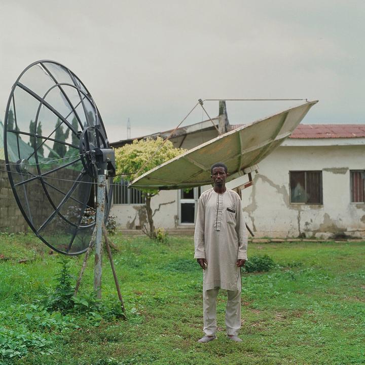 A man stands in a grassy yard in front of a building with two large satellite dishes.