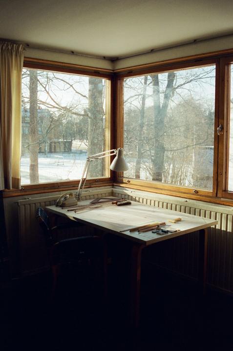 A drafting table with a lamp and tools sits in a corner by a large window overlooking a snowy landscape.
