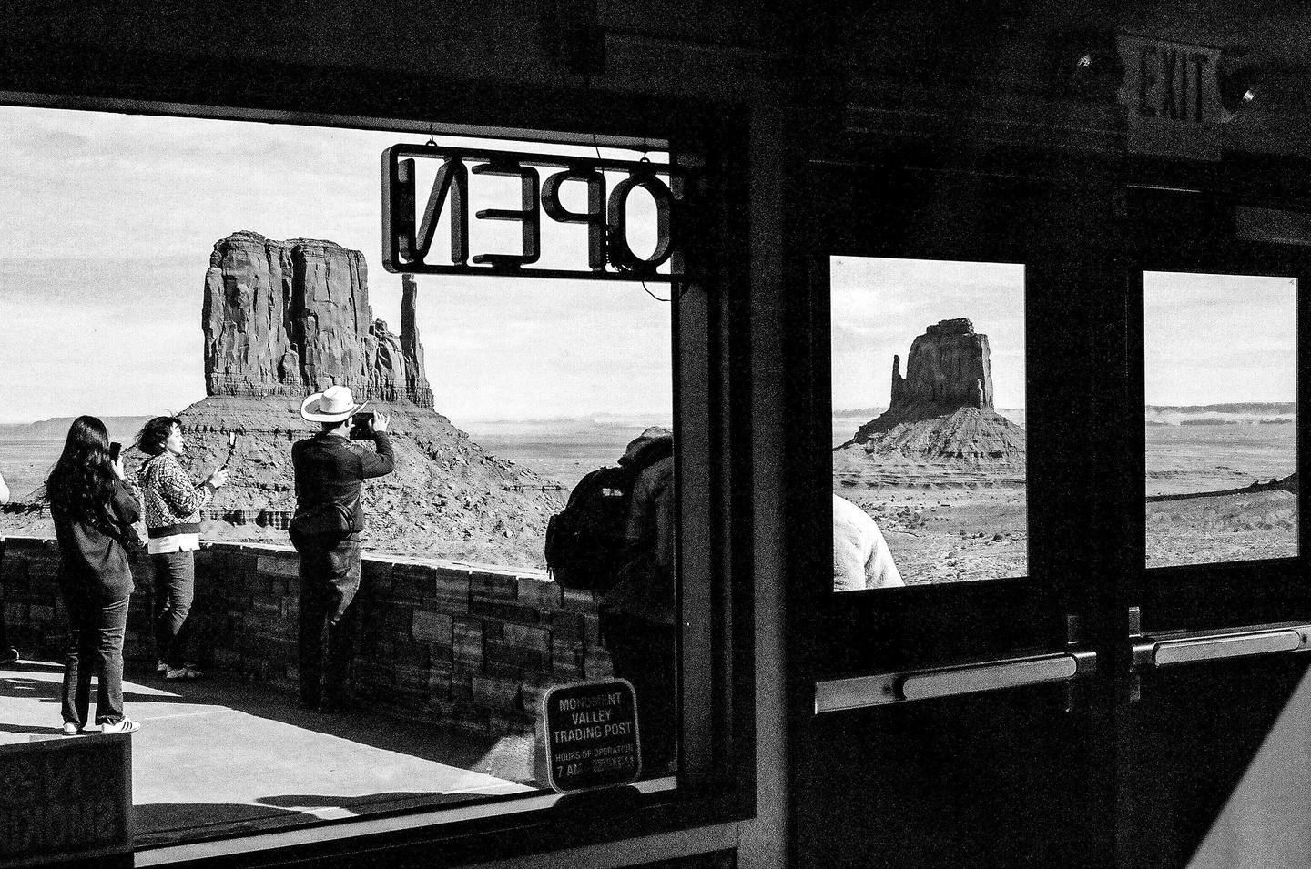 People stand on a balcony overlooking Monument Valley, with a large rock formation visible through a window and an "OPEN" sign reflected in the glass.