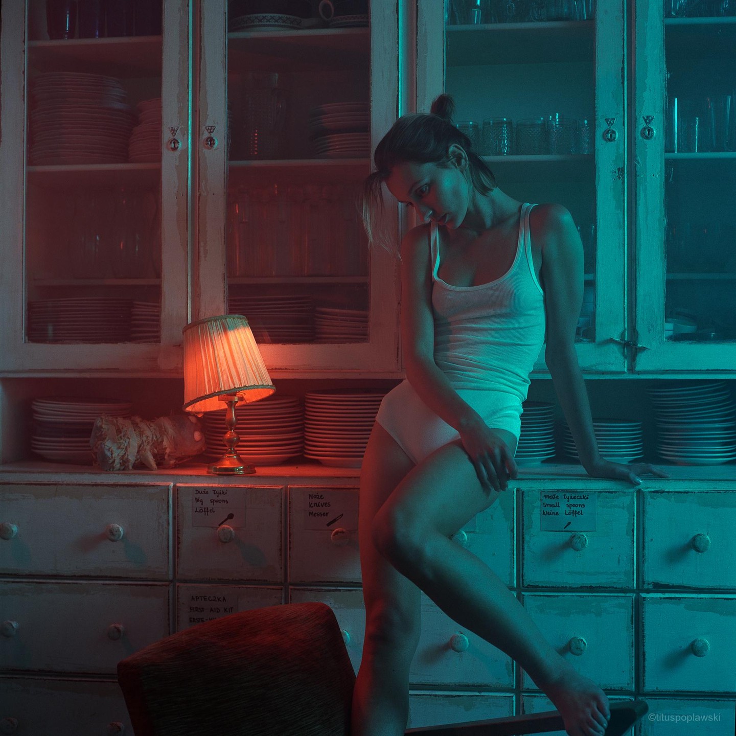 A woman in a white tank top and underwear sits on a cabinet next to a lit lamp.