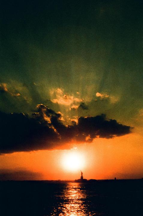 The Statue of Liberty is silhouetted against a bright orange sunset over water with clouds in the sky.