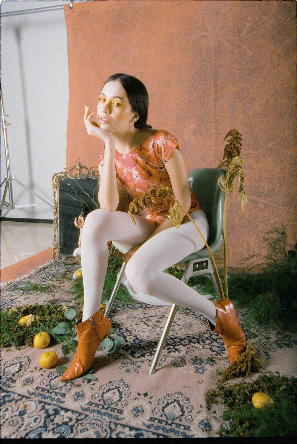A woman with yellow eyeshadow sits on a chair in a studio setting with a patterned rug, plants, and fruit on the floor.