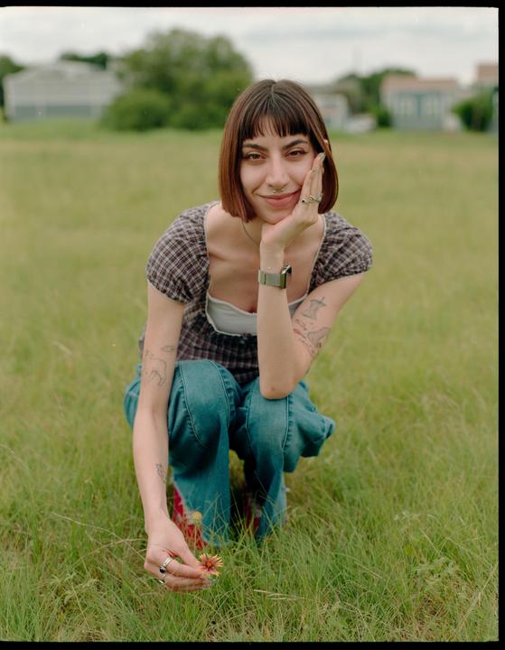 A woman crouches in a grassy field, holding a small flower and looking at the viewer.