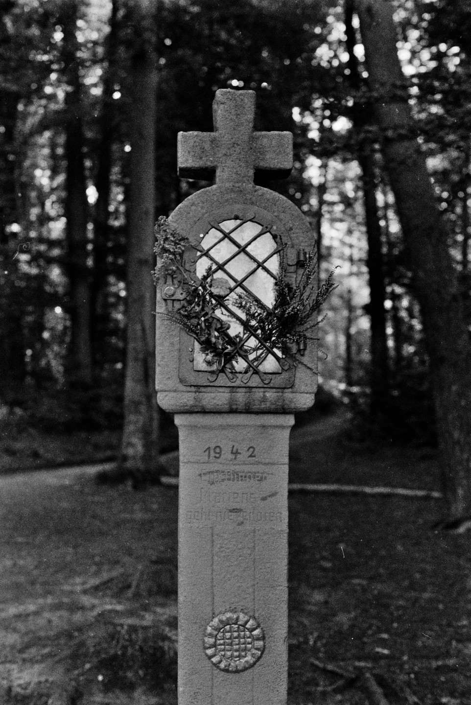 A stone cross with a small, gridded window and floral decorations stands in a forest.
