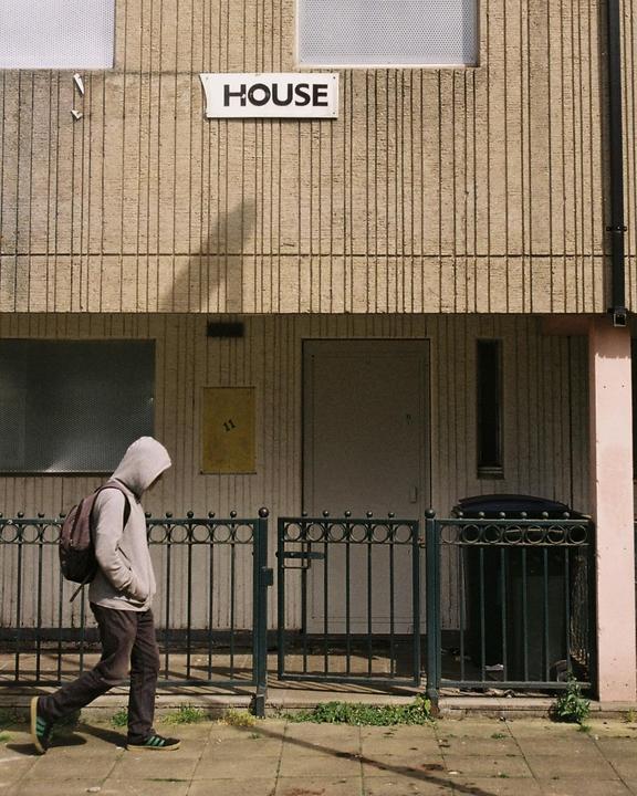 A person wearing a hoodie and backpack walks past a building with a sign that says "HOUSE" on it.