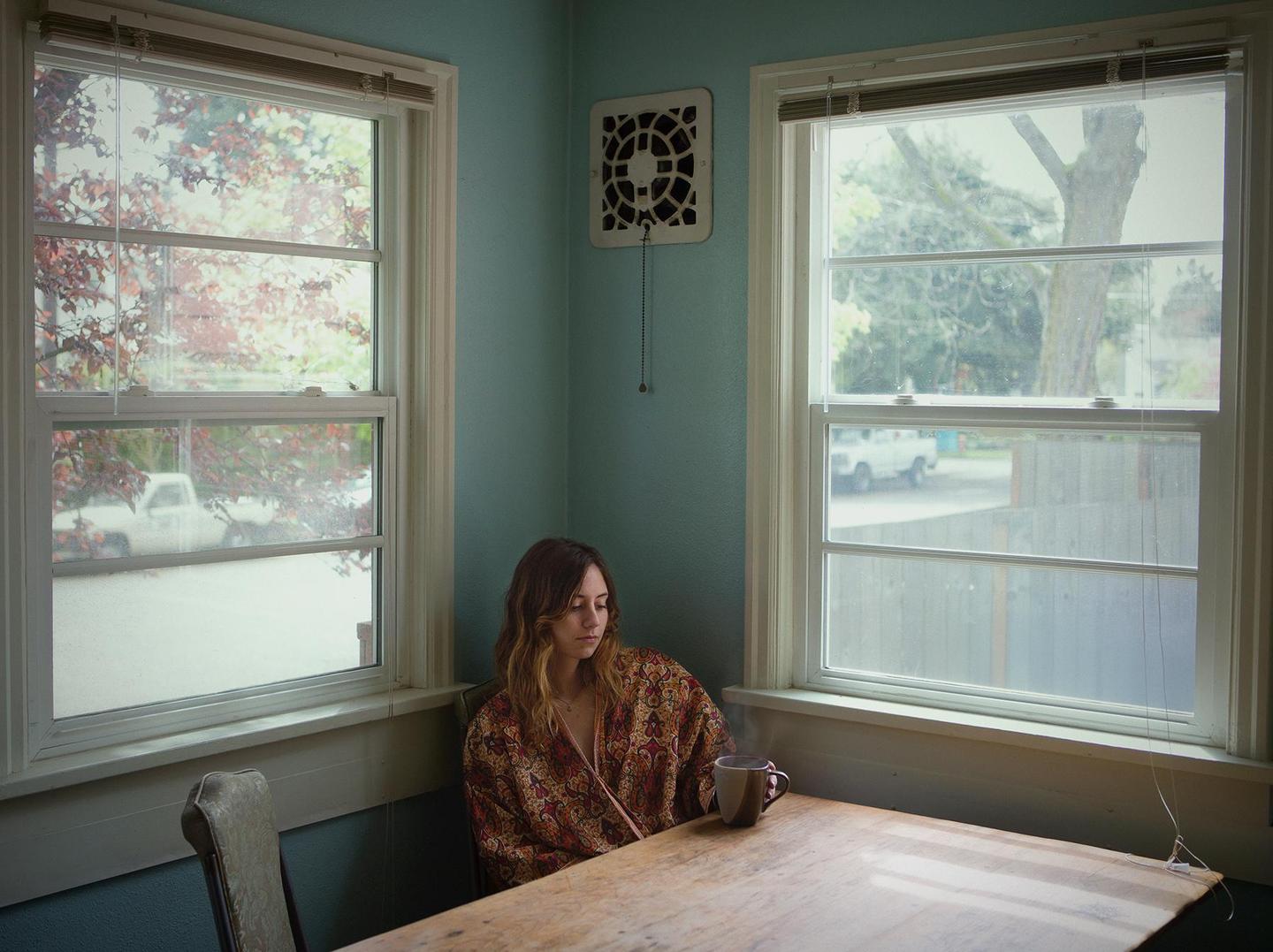 A woman in a patterned kimono sits at a wooden table with a steaming mug between two windows.