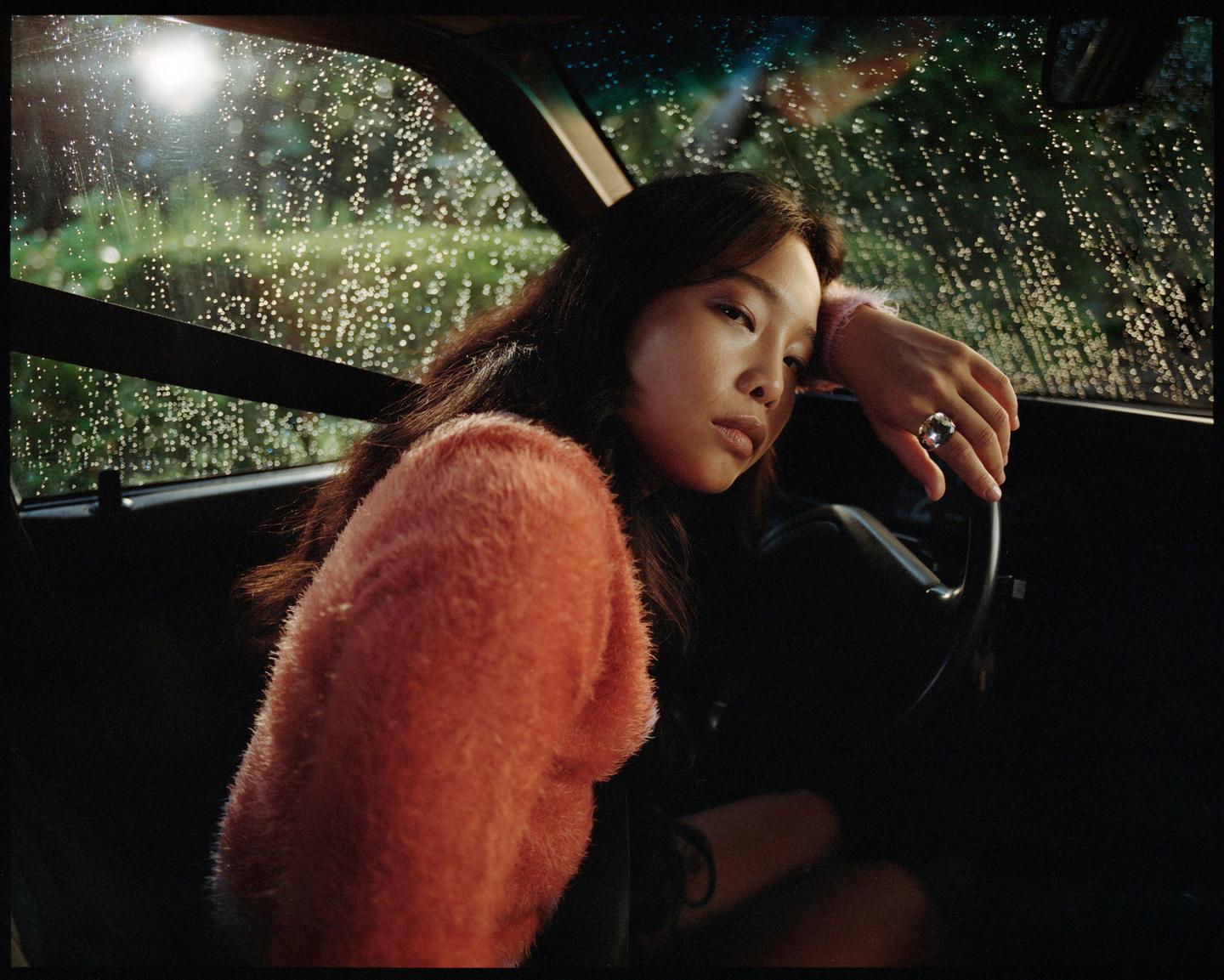 A woman sits in a car with rain on the window.