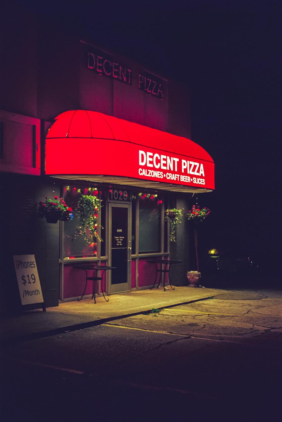 A red awning with the words "Decent Pizza" is illuminated at night above the entrance to a pizza shop.