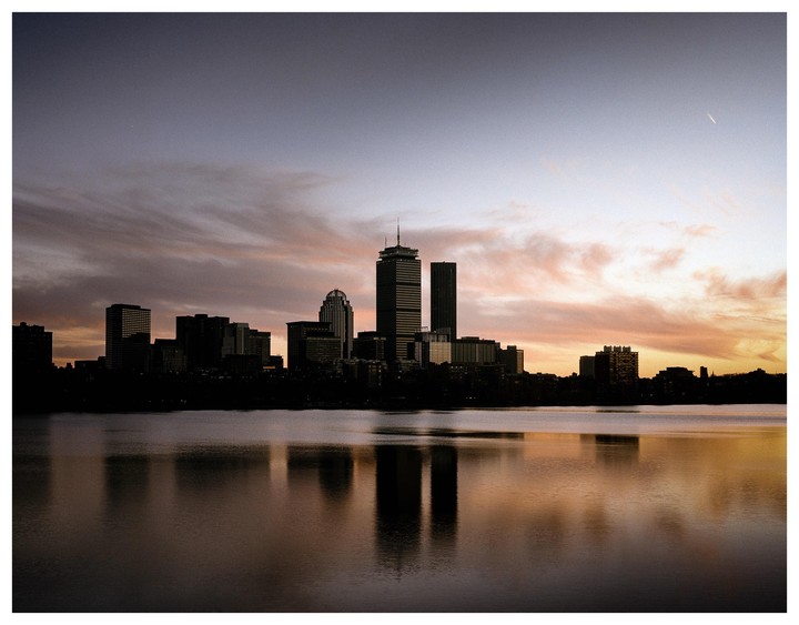 A city skyline is silhouetted against a colorful sunset sky, with reflections visible in the water below.