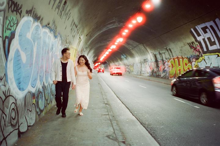 A couple walks hand in hand along a graffiti-covered tunnel wall with cars driving past.