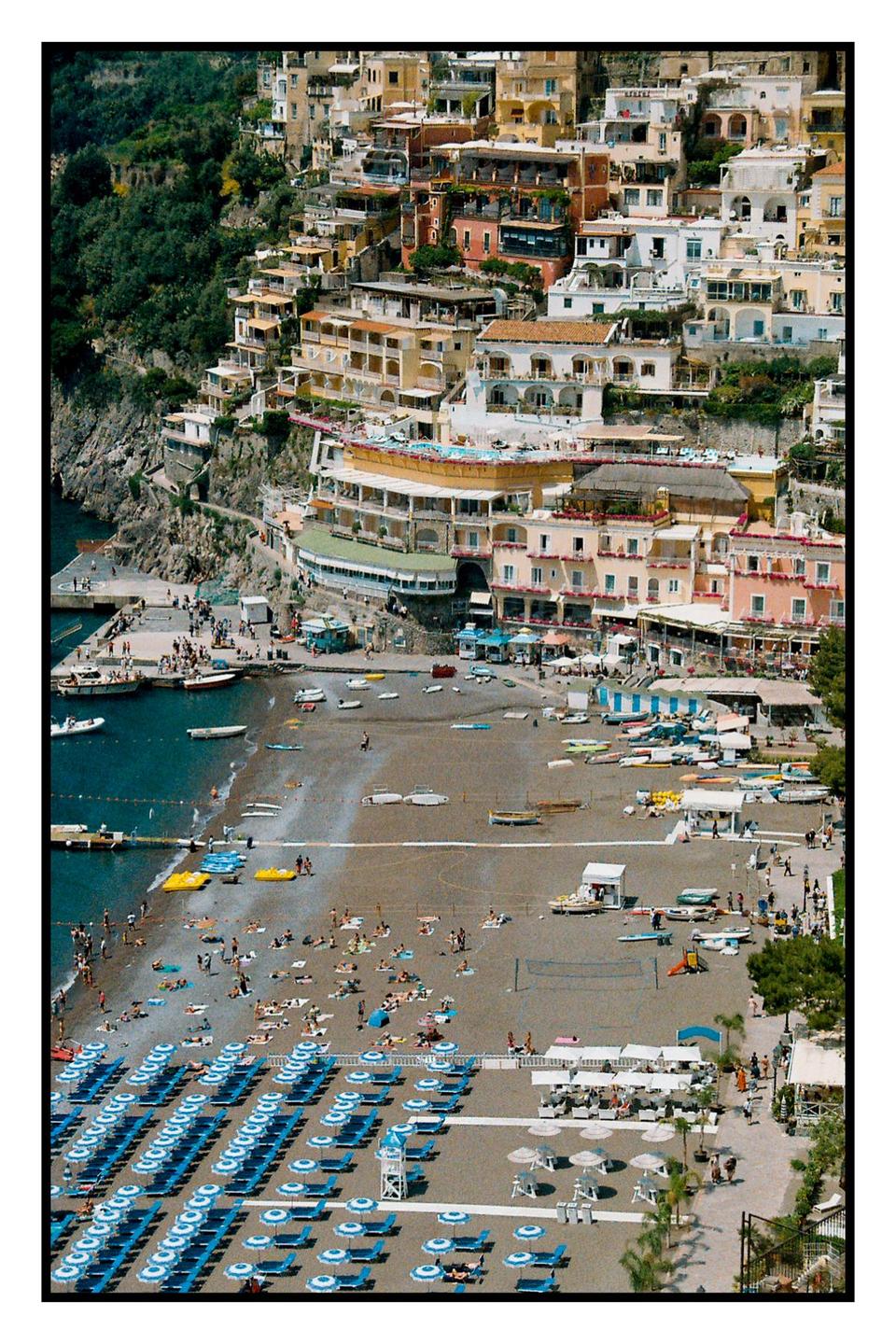 A hillside town with many buildings overlooks a beach filled with people and umbrellas.