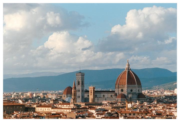 A panoramic view of Florence shows the Duomo and other buildings under a cloudy sky with mountains in the distance.