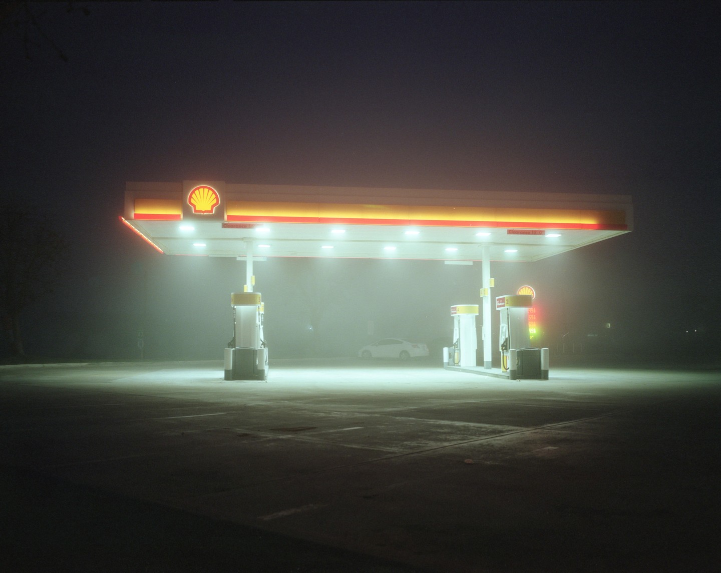 A gas station with a white car is illuminated at night in a thick fog.