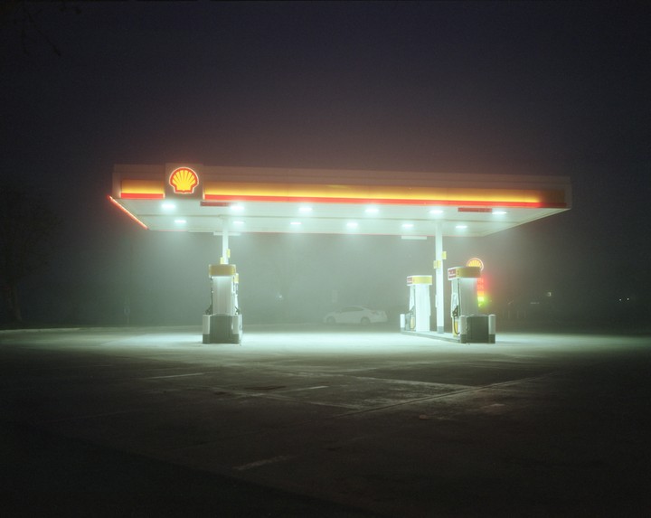 A gas station with a white car is illuminated at night in a thick fog.