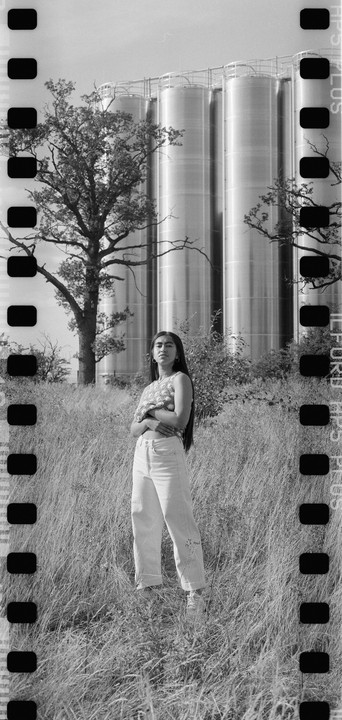 A woman stands in a field of tall grass with large industrial silos and a tree in the background.