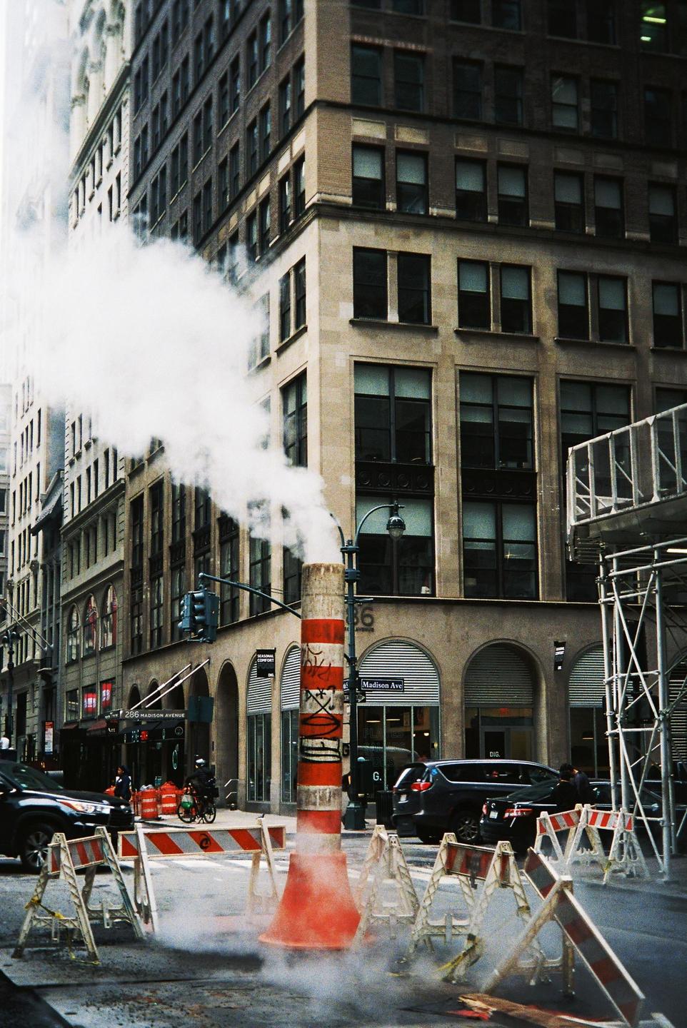 Steam rises from a red and white striped pipe on a city street with tall buildings and cars.