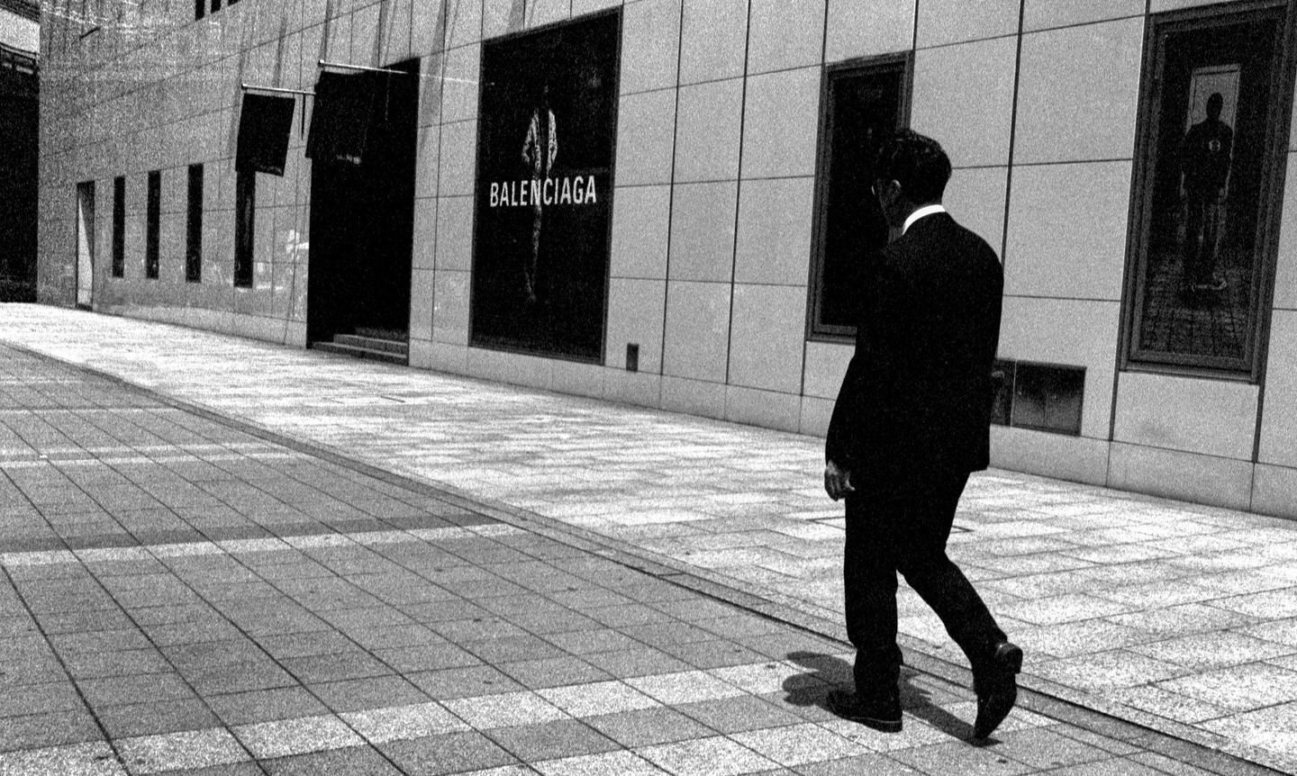 A man in a suit walks past a building with a Balenciaga sign on a sunny day.
